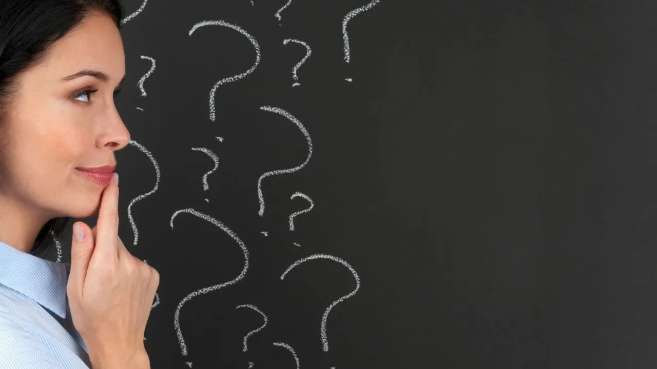 A woman stands in profile with her finger on her chin, appearing thoughtful. She is facing a blackboard covered in chalk-drawn question marks, suggesting she is pondering or considering something.