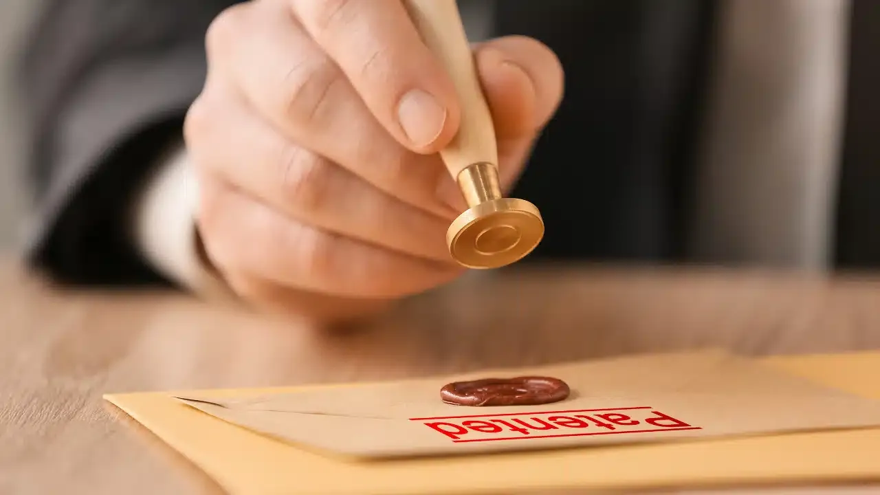 A person presses a brass seal onto a wax seal on a beige envelope labeled Patented in red text, suggesting the official approval of a patent.