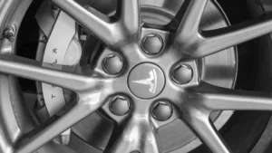 Close-up of a Tesla car wheel, showing the silver alloy rim, lug nuts, and the Tesla logo at the center, with the brake caliper and disc visible in the background.