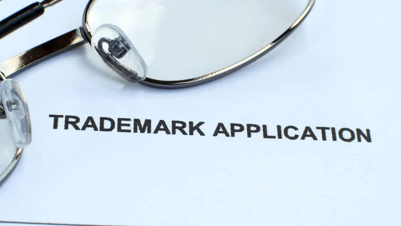 A close-up of a document labeled TRADEMARK APPLICATION in bold black letters, with a pair of eyeglasses resting on the top left corner of the paper.