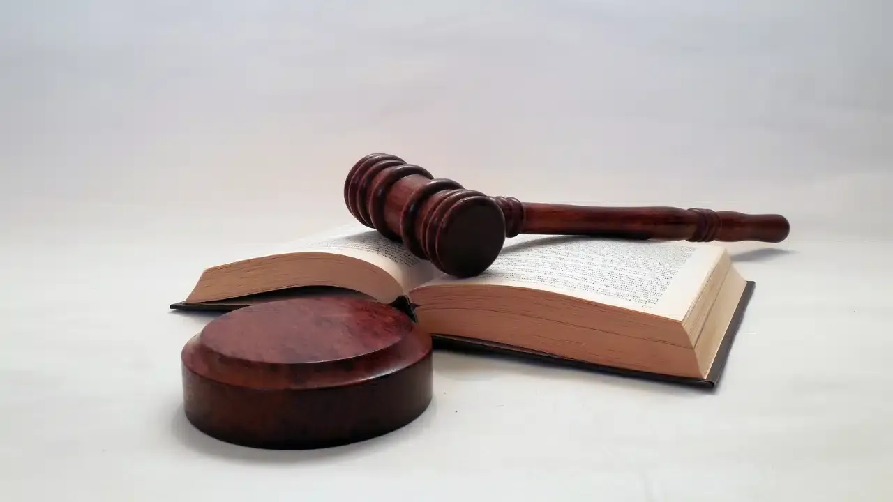 A wooden judge’s gavel rests on an open book, with a round sound block beside them, all placed on a white surface with a plain background.