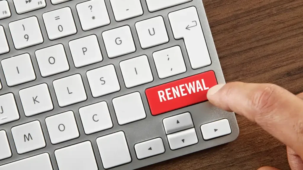 A close-up of a keyboard with a finger about to press a bright red key labeled RENEWAL where the Enter key would normally be. The keyboard is on a wooden desk.