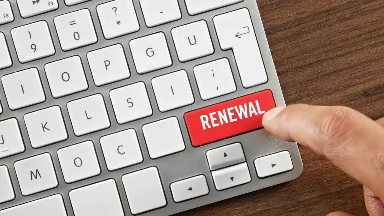 A close-up of a keyboard with a finger about to press a bright red key labeled RENEWAL where the Enter key would normally be. The keyboard is on a wooden desk.