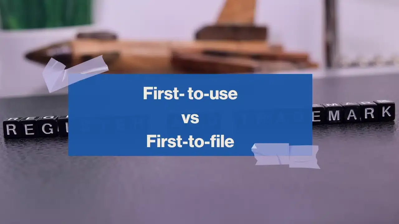 A blue box with white text that reads First-to-use vs First-to-file is overlaid on black letter blocks spelling out REGISTER AND TRADEMARK on a dark surface, with blurred objects in the background.