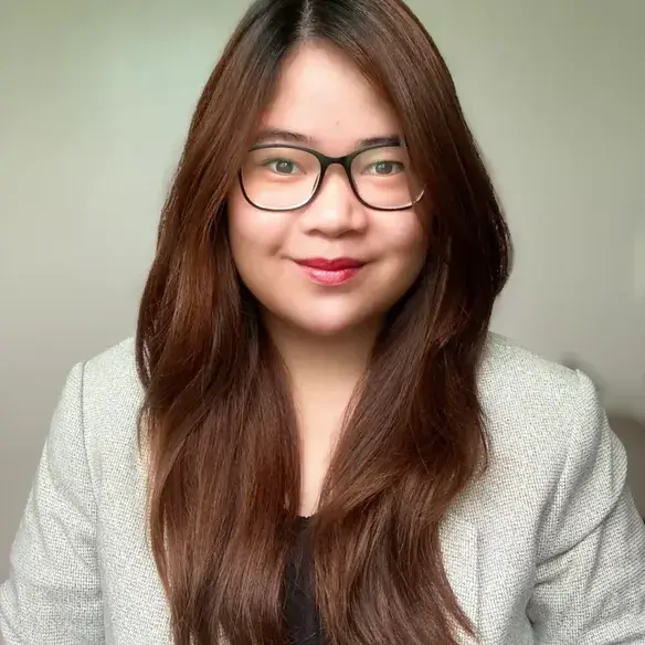 A woman with long brown hair and glasses, wearing a light gray blazer and black top, is smiling at the camera against a plain light background.