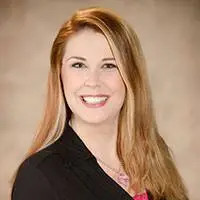 A woman with long light brown hair, wearing a black blazer and a pink top, smiling at the camera against a plain beige background.
