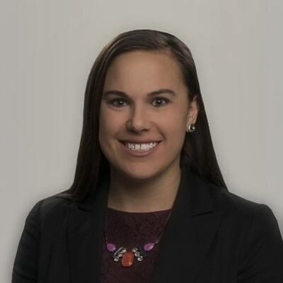 Woman with long dark hair, wearing a black blazer over a burgundy patterned top and statement necklace, smiles at the camera against a neutral gray background.