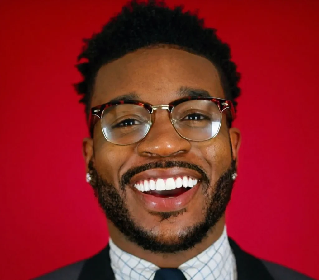 A man with short, curly hair, beard, glasses, and earrings smiles widely, wearing a suit and tie, in front of a bright red background.