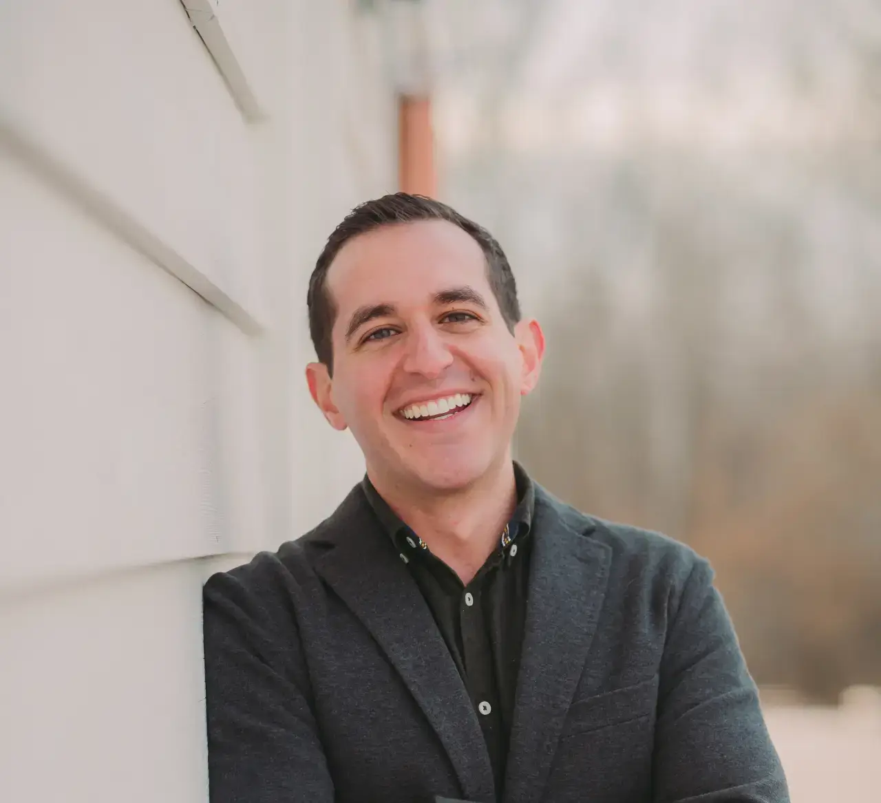 A man with short dark hair, wearing a dark blazer and black shirt, stands outdoors leaning against a wall, smiling with arms crossed.
