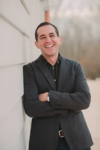 A man in a dark blazer stands outdoors, leaning against a light-colored wall with arms crossed, smiling at the camera.