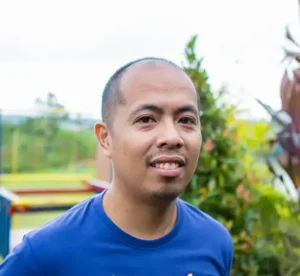 A man with a shaved head and a short mustache and goatee, wearing a blue shirt, stands outdoors with greenery in the background.