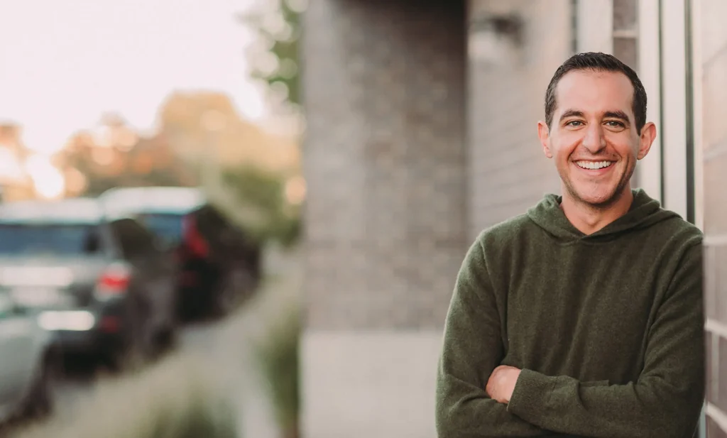 A man in a green sweater stands outside with arms crossed, smiling at the camera. A brick wall is behind him, and blurred cars and trees are in the background.