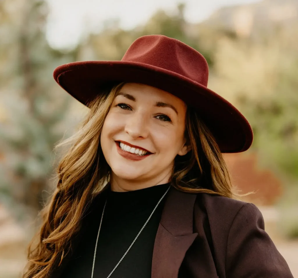 Woman with long brown hair, wearing a wide-brimmed burgundy hat and matching blazer, smiles outdoors with blurred greenery in the background.