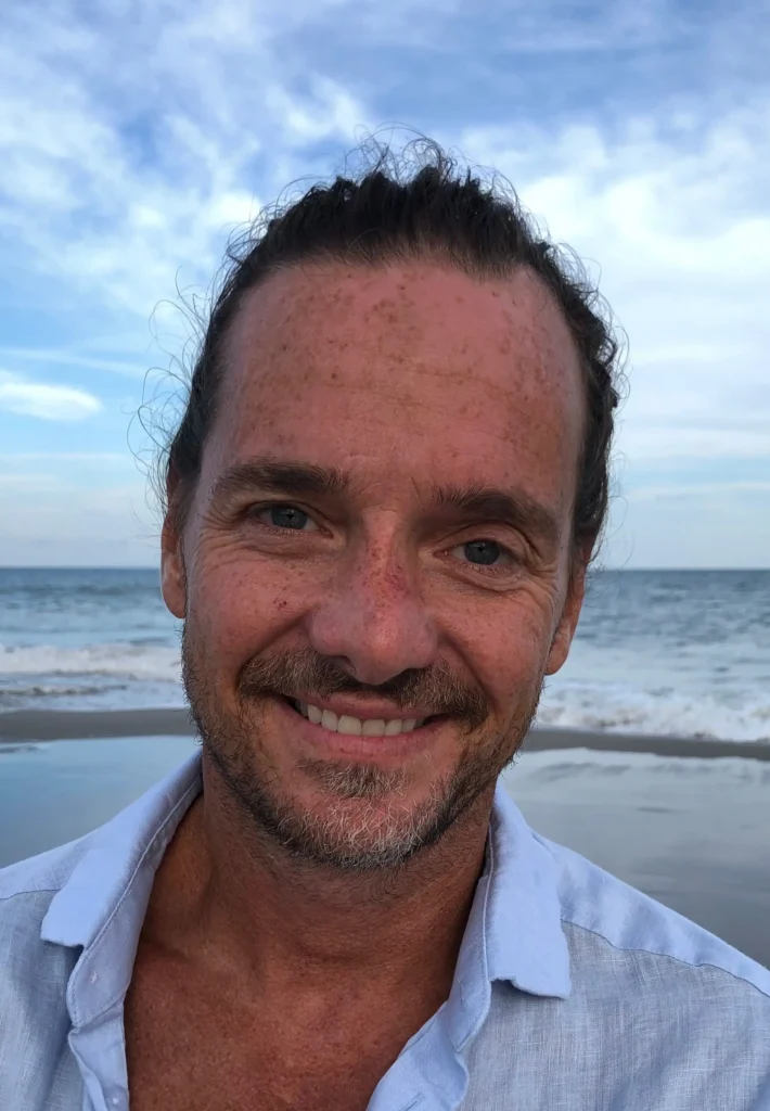 A man with a light beard and mustache smiles at the camera on a beach with waves and a cloudy sky in the background.