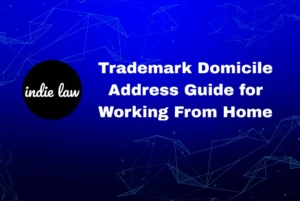 Blue graphic with white text reading "Trademark Domicile Address Guide for Working From Home" and a black circle logo with "indie law" in white. Geometric line patterns highlight the focus on Trademark Domicile Address rules.