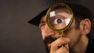 A man wearing a pinstripe fedora holds a magnifying glass up to one eye, making his eye appear much larger. He has a beard and mustache, and is looking directly at the camera against a plain background.