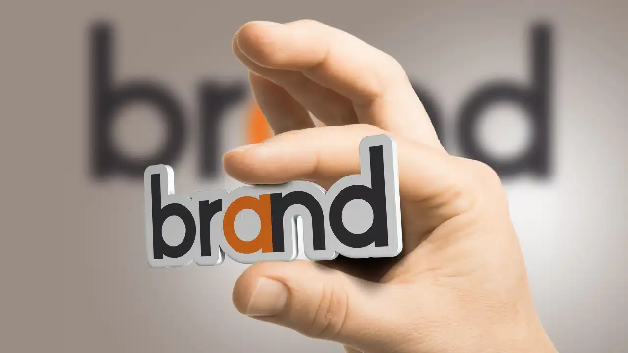 A hand holds a cutout of the word brand in bold letters, with the background showing a blurred, larger version of the same word. The a in brand is highlighted in orange.