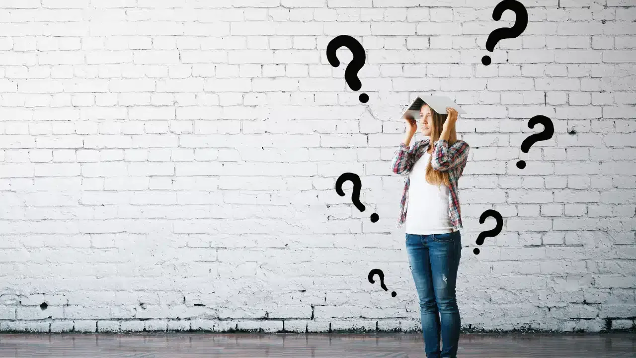 A person stands against a white brick wall holding a book on their head, looking confused. Large black question marks are drawn around them, emphasizing uncertainty or confusion.