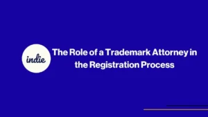 A blue slide with a white circle on the left containing the word indie in cursive, and the text The Role of a Trademark Attorney in the Registration Process in bold white letters. Thin yellow lines are at the bottom right.