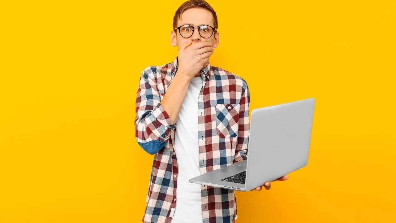 A surprised person wearing glasses and a plaid shirt holds a laptop in one hand and covers their mouth with the other, standing against a bright yellow background.