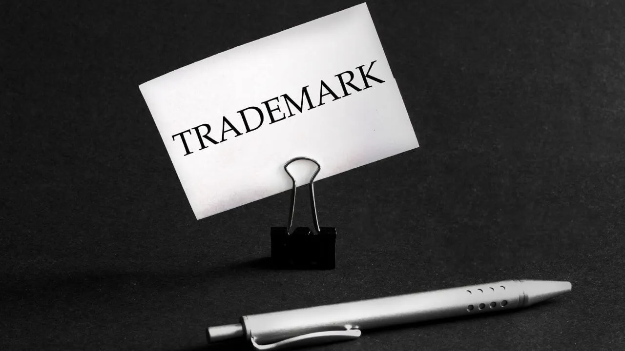 A card with the word TRADEMARK clipped to a black binder clip stands on a dark surface, with a silver pen lying in the foreground.
