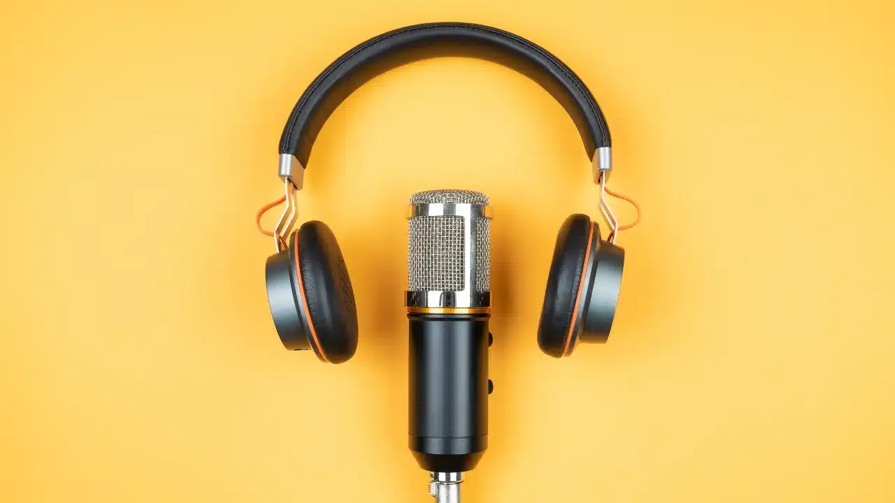 A pair of black headphones surrounds a silver microphone, both placed on a bright yellow background. The headphones form a semi-circle above the microphone, creating a visually balanced composition.