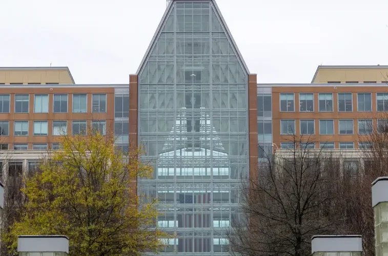 A modern office building with a large glass facade, reflecting an interior atrium, flanked by trees with sparse and yellowing leaves.