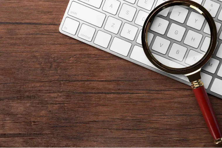 A magnifying glass resting on a computer keyboard placed on a wooden surface.
