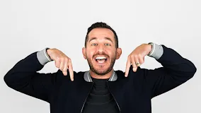 A smiling man with short dark hair points downward with both hands, wearing a black jacket over a black shirt against a plain white background.