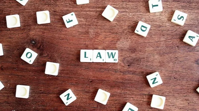 Scrabble tiles on a wooden surface spell out "LAW" in the center, surrounded by scattered, unused letter tiles.
