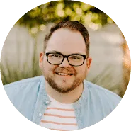 A smiling man with short hair, glasses, a beard, and mustache, wearing a light blue shirt over a white and pink striped shirt, stands outdoors with greenery in the background, exuding confidence like someone well-versed in trademarks.