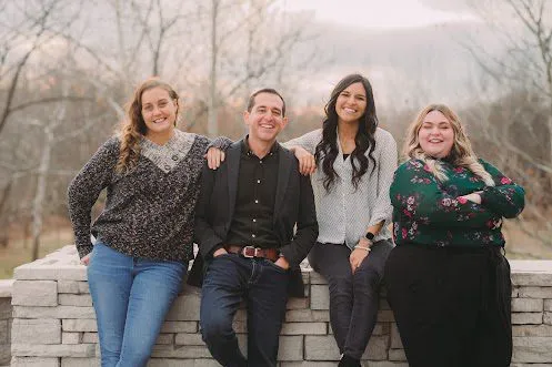 Four people are posing together outdoors, smiling, and leaning against a low stone wall. The background features bare trees, suggesting its autumn or early winter.