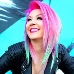 Smiling person with vibrant pink and pastel green hair, wearing a black leather jacket, poses in front of a bright blue background—showcasing their unique style as their own personal trademark.