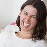 A woman with long dark hair and a white shirt is sitting indoors, smiling widely and laughing, looking slightly to the side. Light background with pink and white paper partially visible behind her, adding a playful touch reminiscent of creative trademarks.