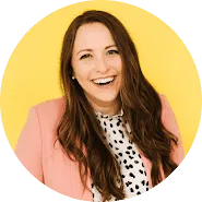 A woman with long brown hair, wearing a pink blazer and a white blouse with black spots, smiles in front of a bright yellow background—a vibrant look that stands out like unique trademarks in fashion.