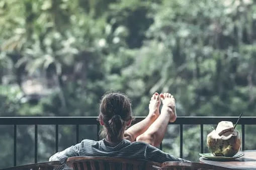 A person relaxes on a balcony with feet up on the railing, looking at lush green trees. A fresh coconut with a straw sits on a table nearby, its label adorned with colorful trademarks.