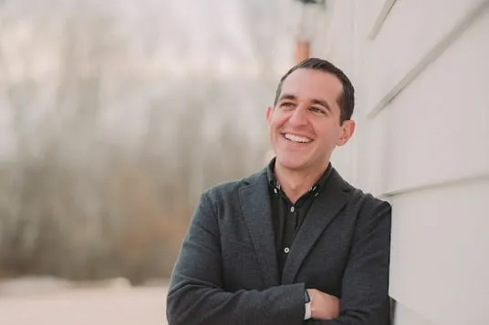 A man in a dark blazer stands outdoors, leaning against a white building with his arms crossed and smiling, with trees blurred in the background.