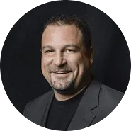 Smiling man with short dark hair and a trimmed beard, wearing a dark blazer over a black shirt, posed in front of a black background, exuding confidence often seen in trademarks of success.