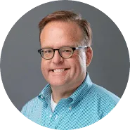 A man with short light brown hair and glasses smiles at the camera. He is wearing a blue and white checkered button-up shirt, perfect for a trademarks professional. The background is plain gray.