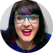 A person with long dark hair, blue glasses, and bright pink lipstick—true trademarks of their style—is smiling widely at the camera. They are wearing a blue top and red tassel earrings.